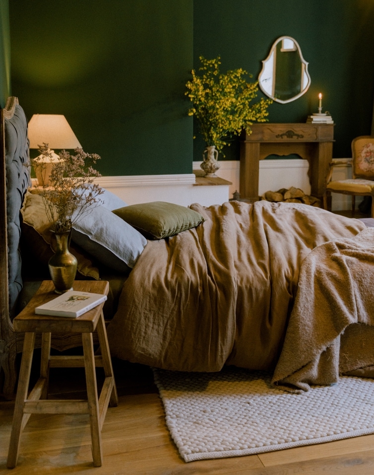 Chambre chaleureuse avec linge de lit en lin marron et coussins verts sur mur vert foncé