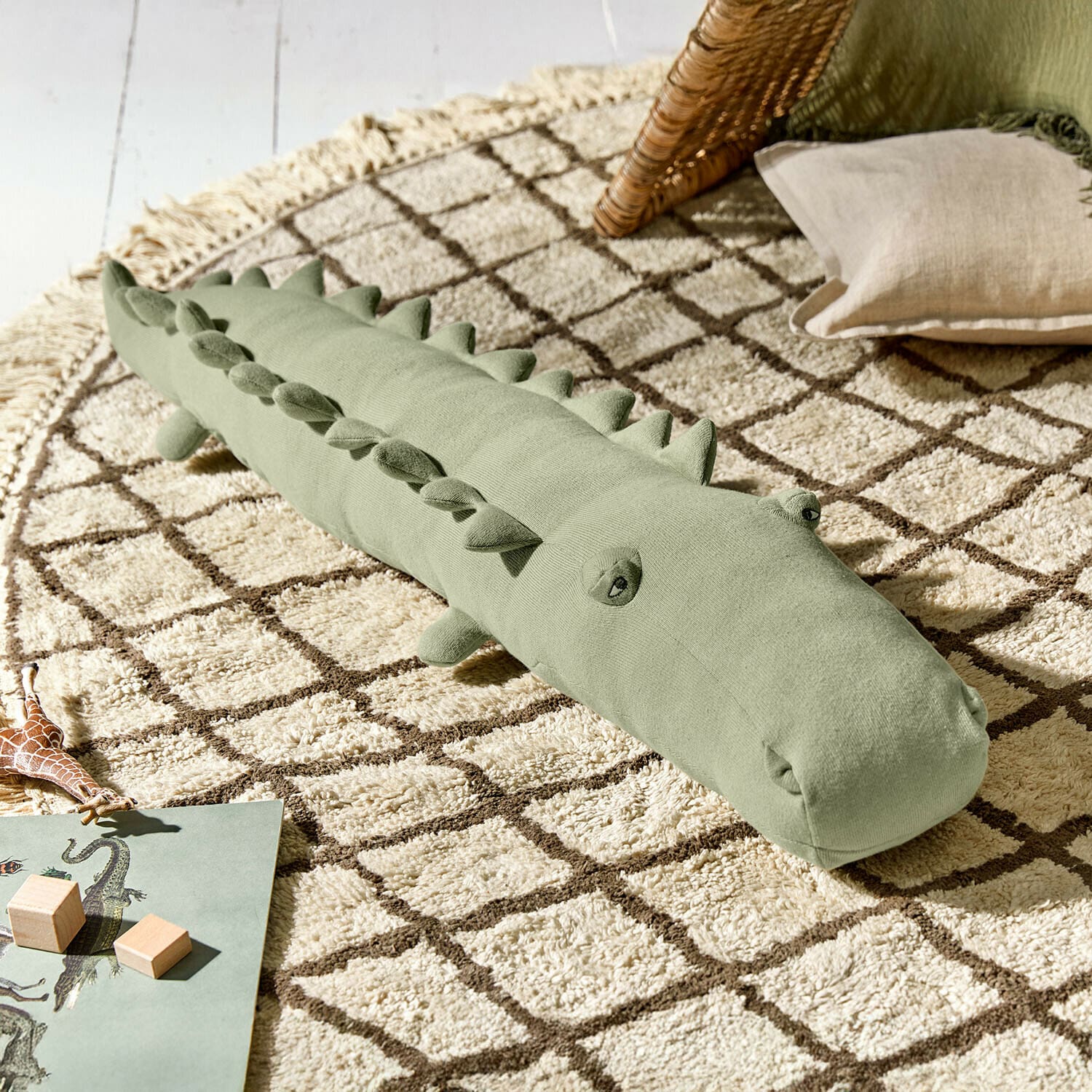 Peluche Zimpy Coussin doux vert en forme de crocodile sur un tapis beige à motif géométrique