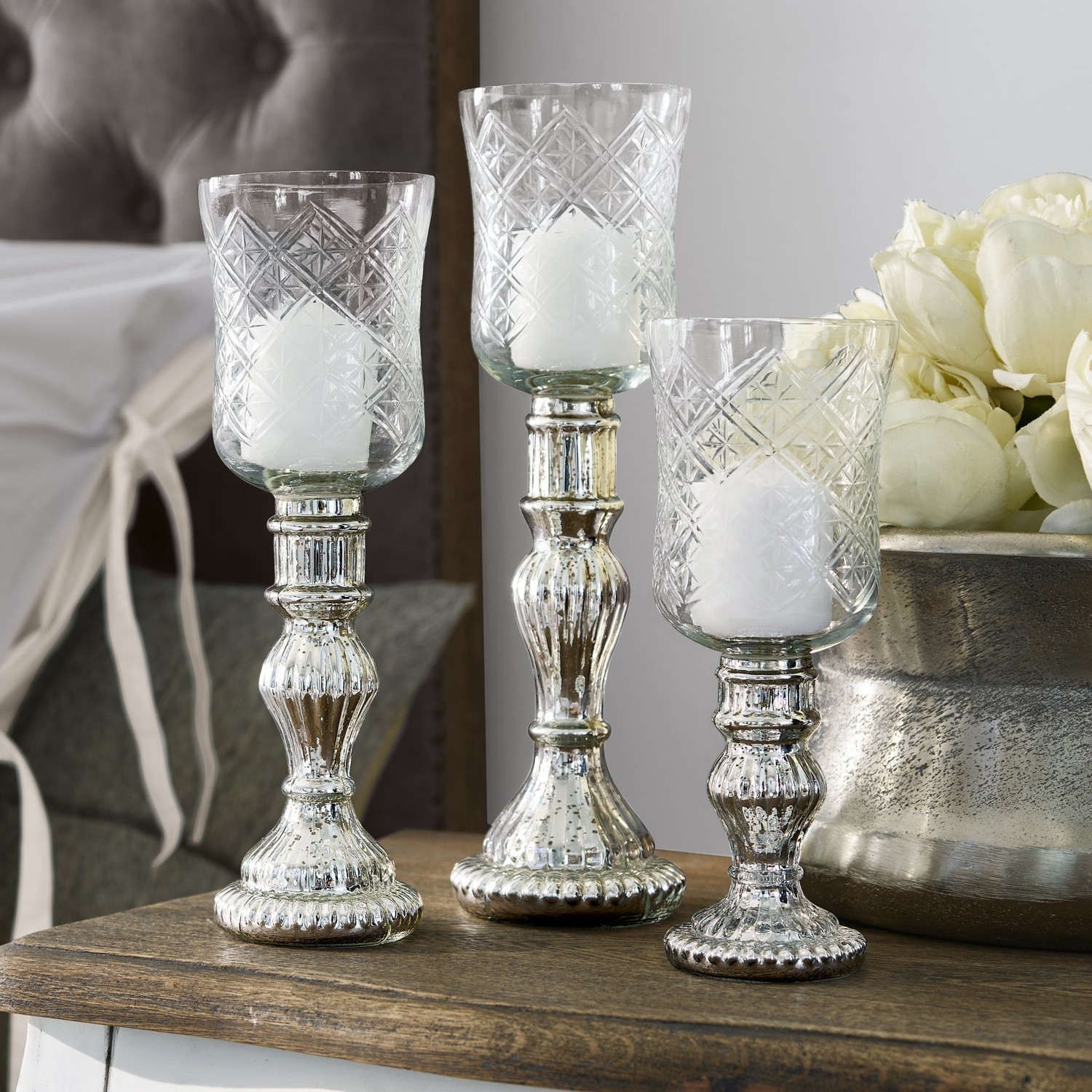 Lot de 3 photophores Celestin Ensemble de trois bougeoirs en verre avec base métallique argentée posés sur une table en bois