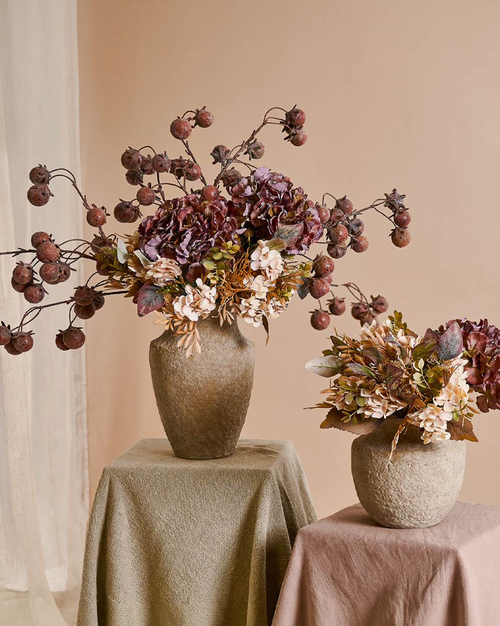 Deux vases décoratifs en pierre beige avec fleurs d'automne et fruits secs sur tables drapées