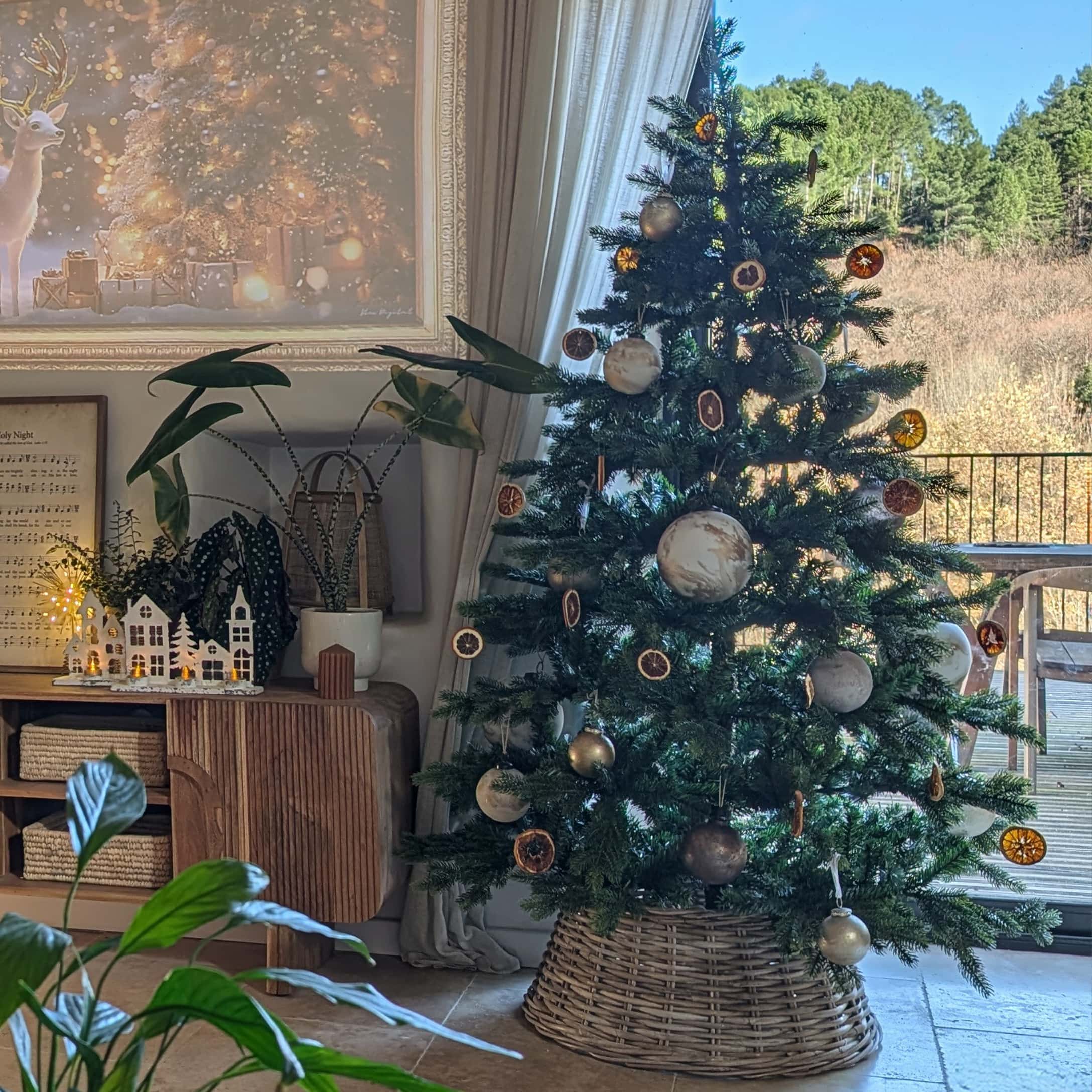 Sapin de Noël décoré avec des ornements naturels et base en osier dans un salon lumineux avec vue sur la forêt