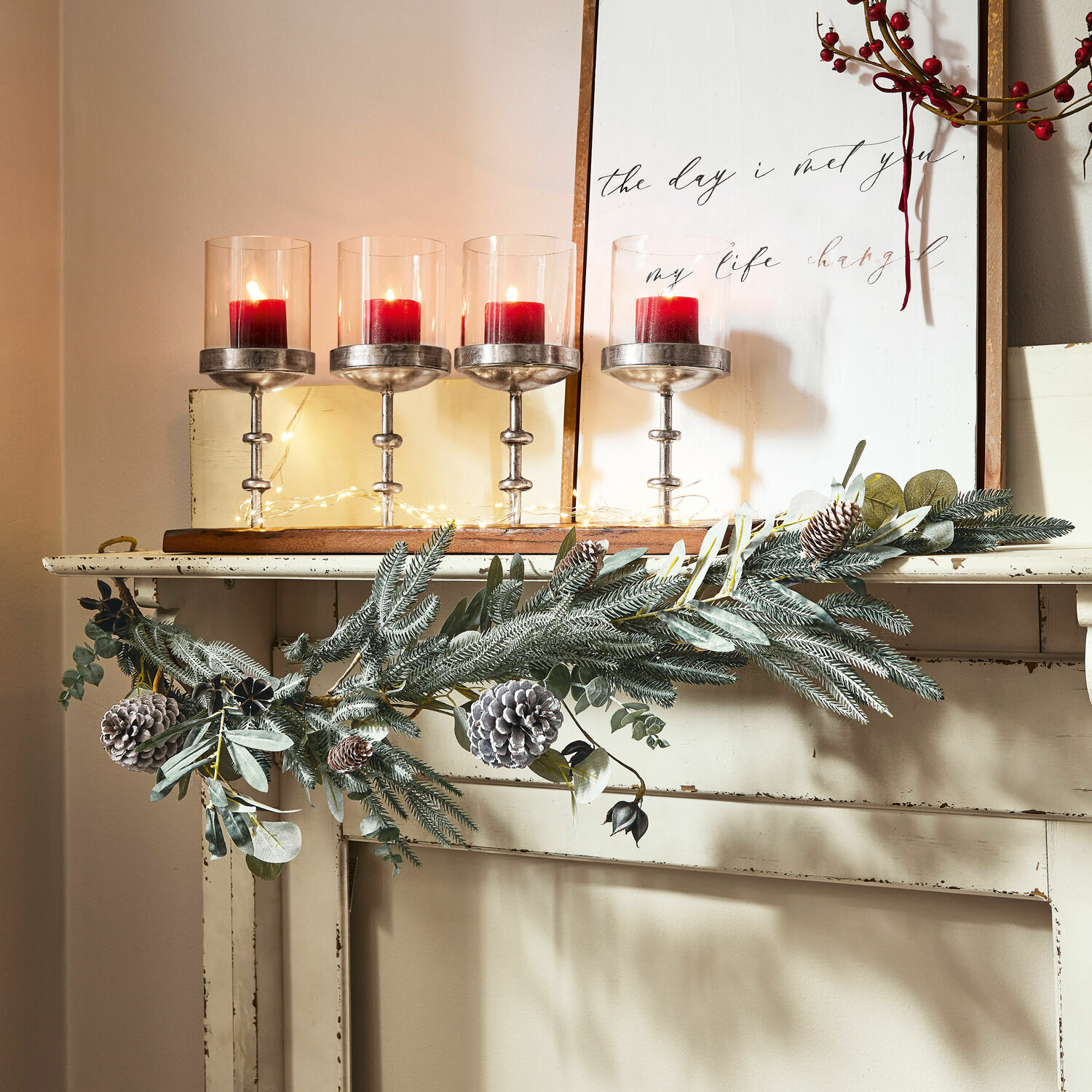 Guirlande Rosella Couronne décorative avec pommes de pin et branches vertes sur une cheminée blanche avec bougies rouges