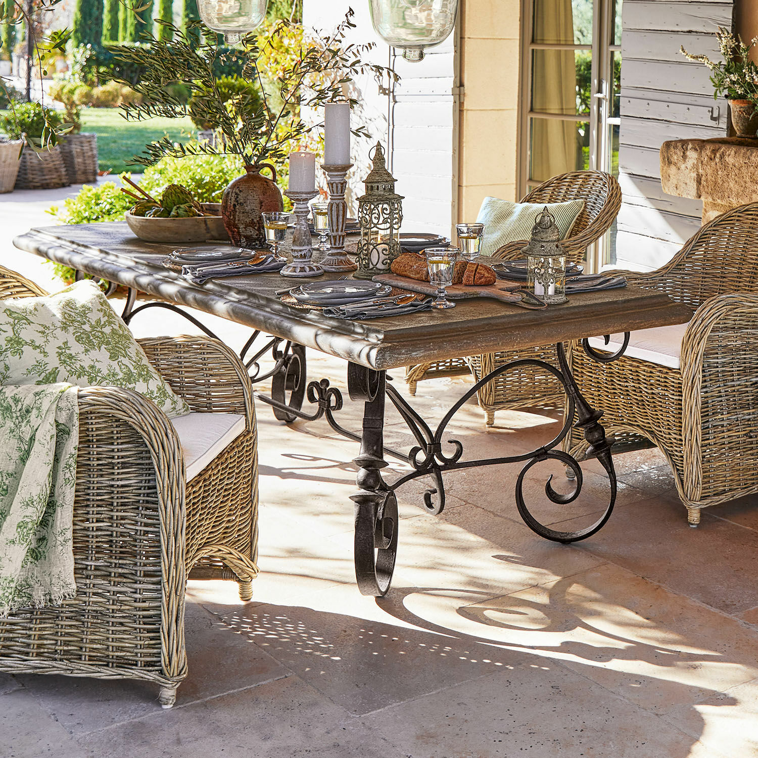 Table Bréhéville Table de jardin avec pieds en métal et plateau en bois, entourée de fauteuils en osier avec coussins