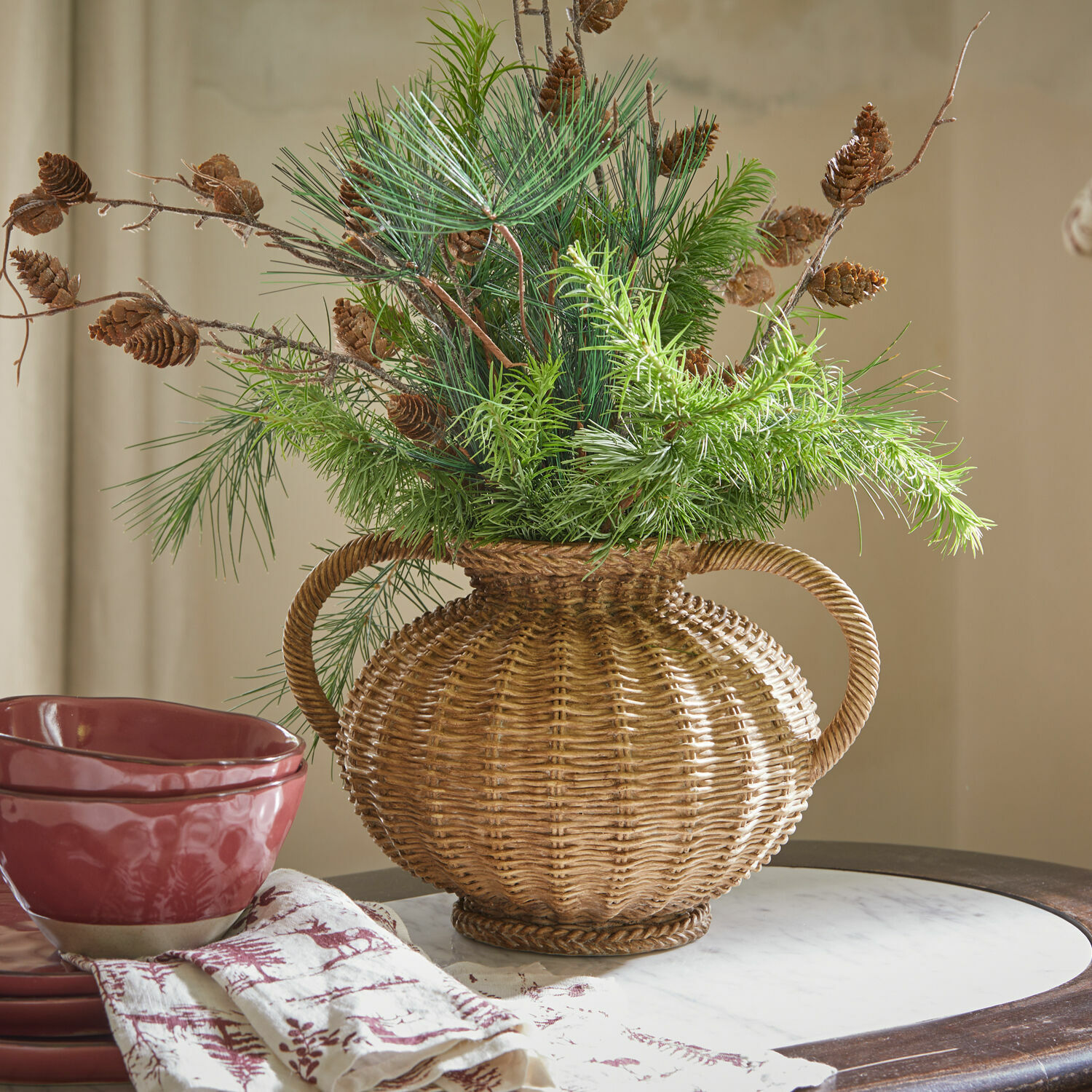 Vase Kaelvik Vase en osier avec poignées, décoré de branches vertes et pommes de pin sur une table avec vaisselle rouge
