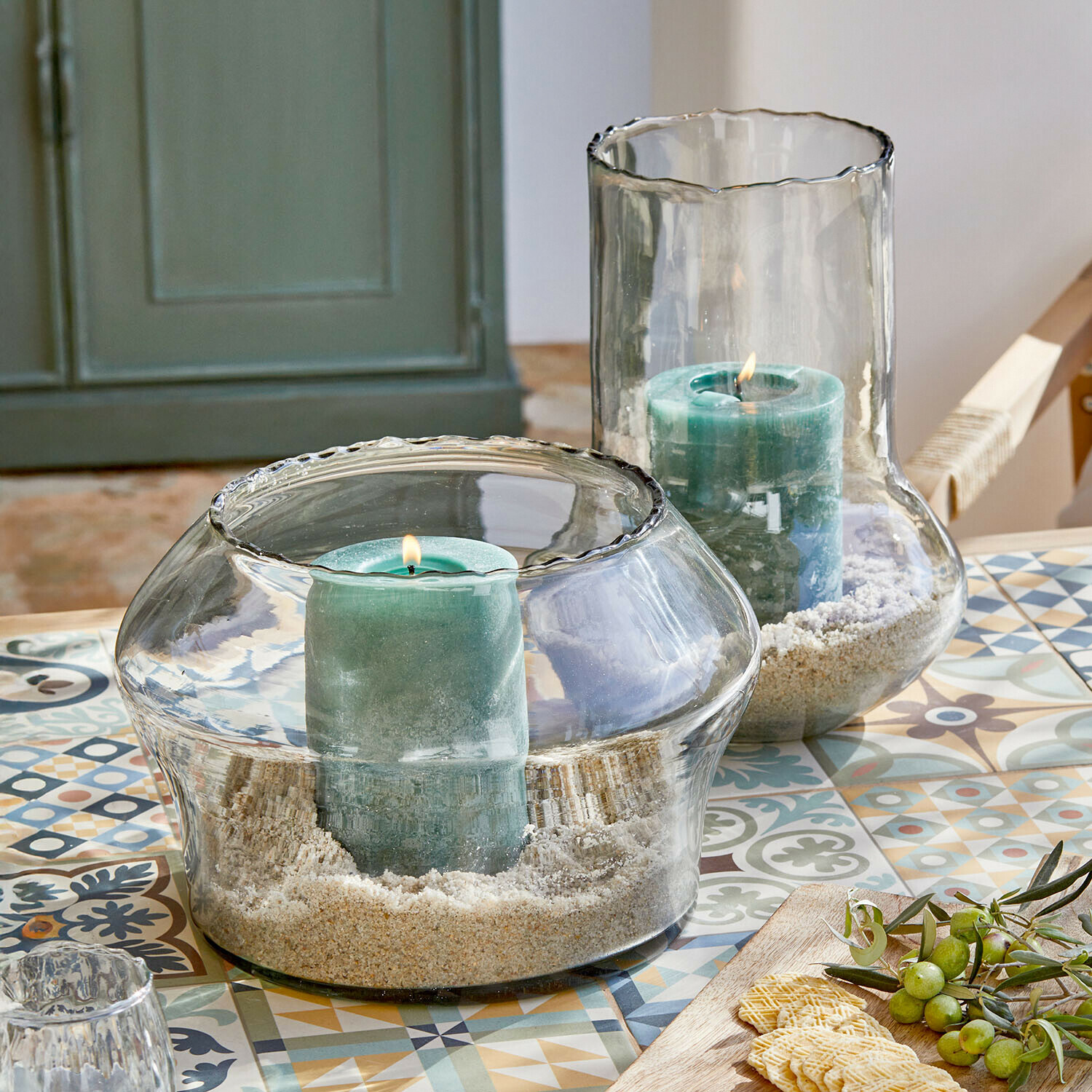 Lot de 2 photophores Lumiras Bougeoirs en verre avec bougies vertes et sable sur une table colorée à motifs décoratifs