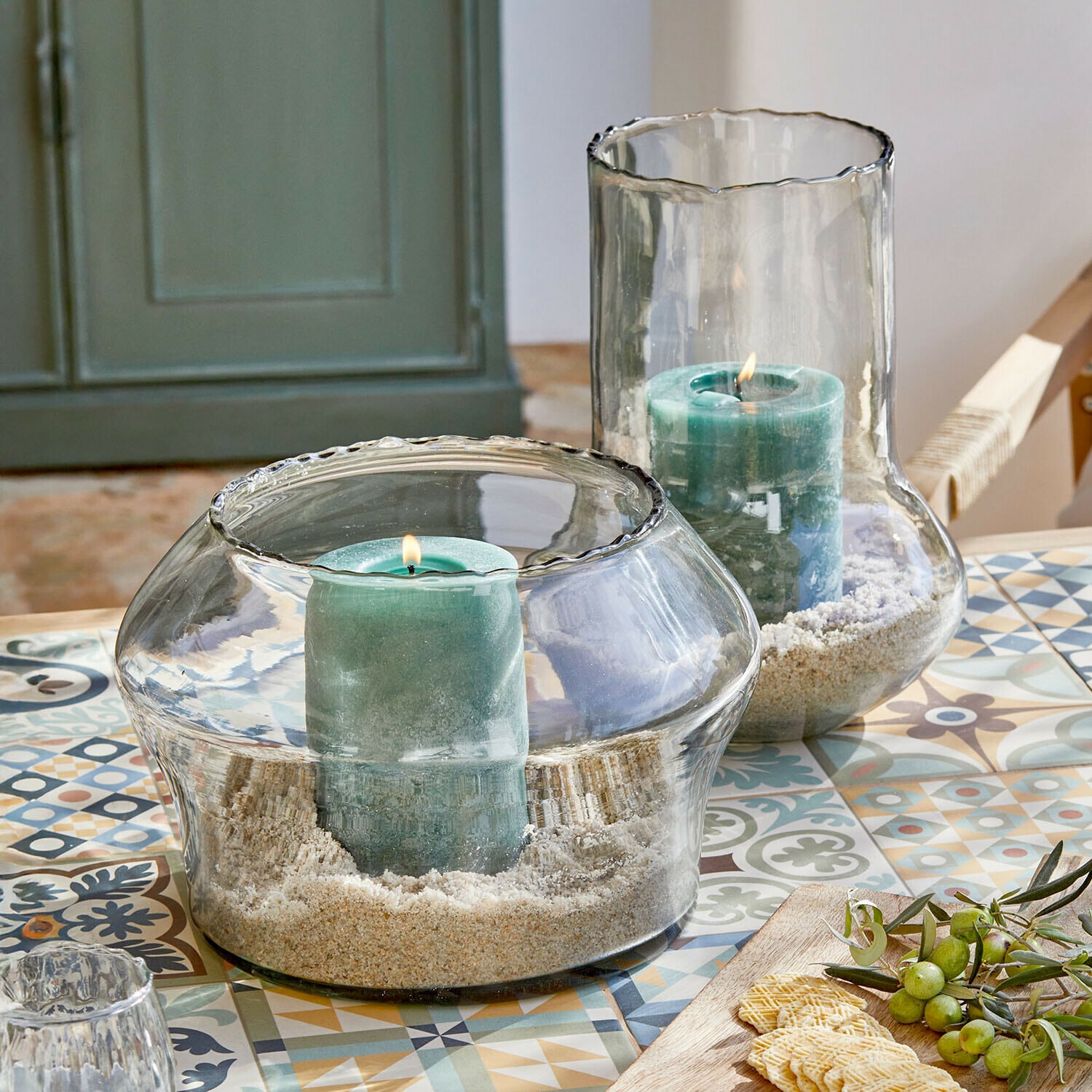 Lot de 2 photophores Lumiras Bougeoirs en verre avec bougies vertes et sable sur une table colorée à motifs décoratifs