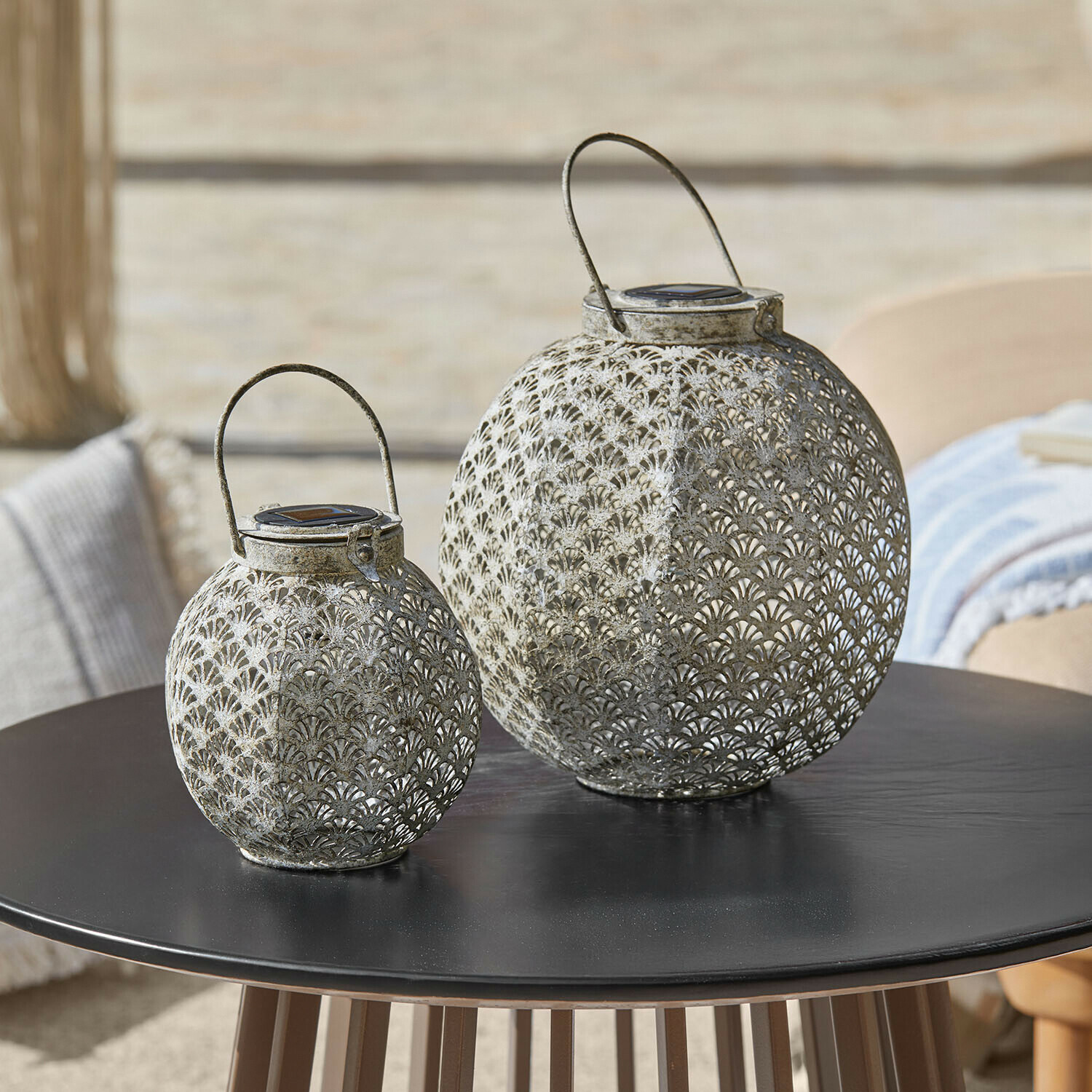 Lot de 2 lanternes solaires Omirah Deux lanternes en métal ajouré gris posées sur une table noire, décoration extérieure élégante