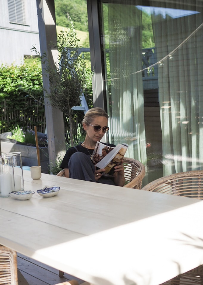 Femme lisant un livre sur une terrasse avec une table en bois clair et des chaises en osier