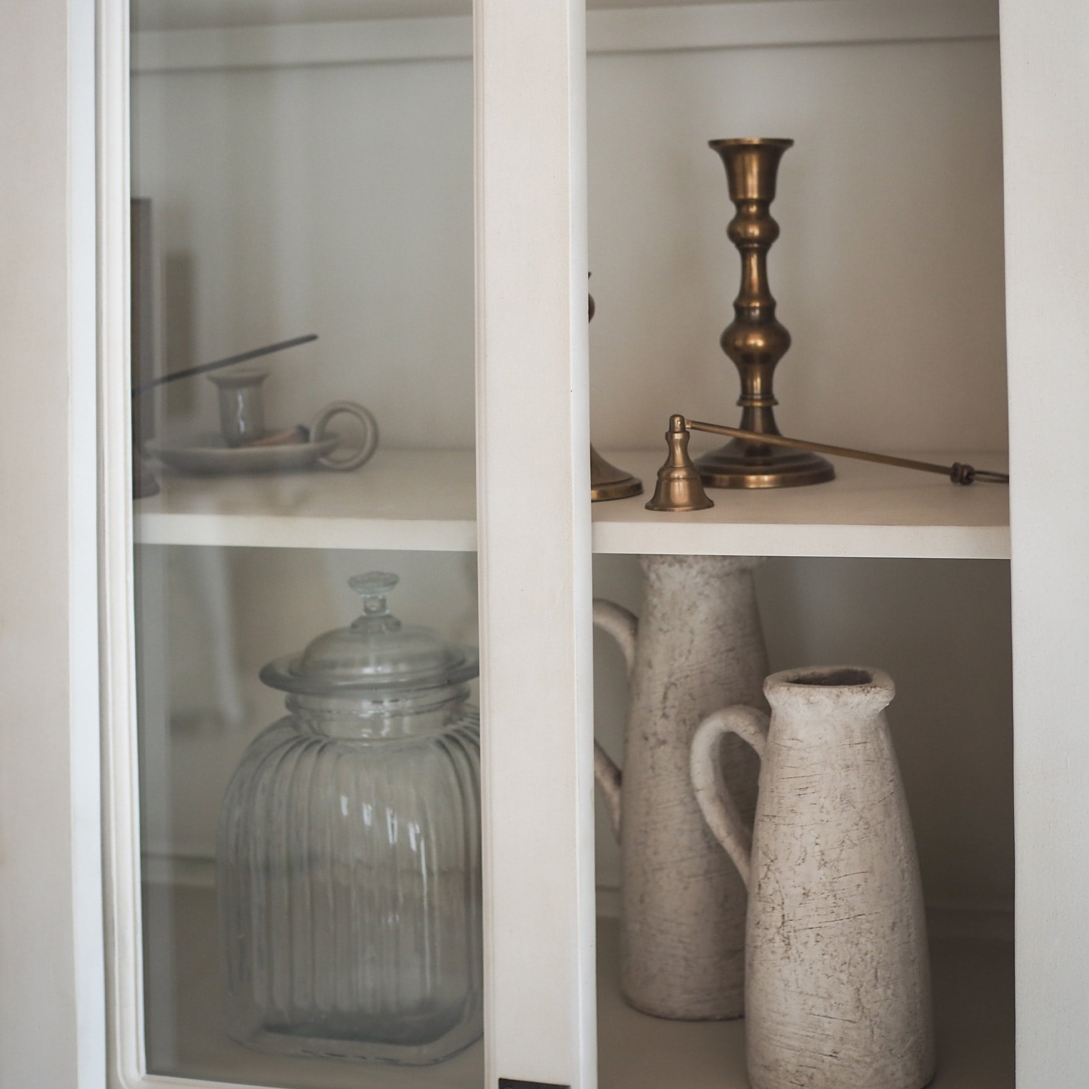 Pot en verre et pichets en céramique blanche dans une armoire avec bougeoir en laiton et clochette