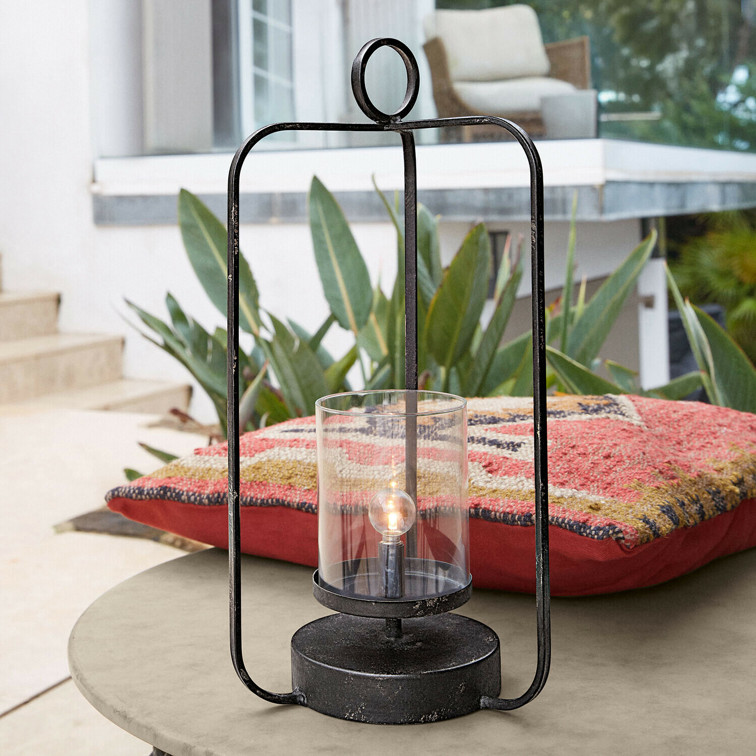 Lanterne LED Kivalex Lanterne noire en métal avec verre transparent posée sur une table de jardin avec coussin coloré