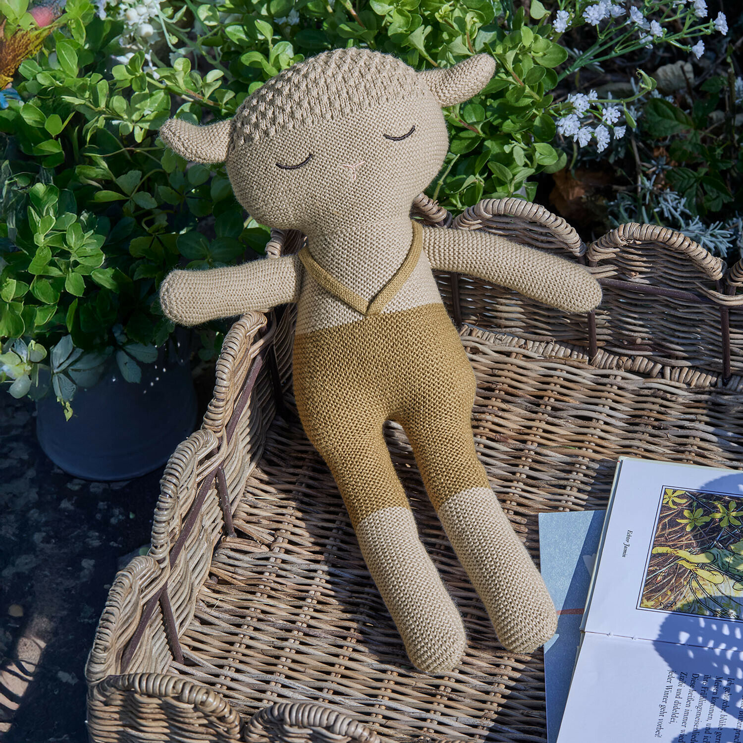 Peluche Cuddlecloud Peluche en forme de mouton beige en tricot posée dans un panier en osier près d'un livre dans un jardin