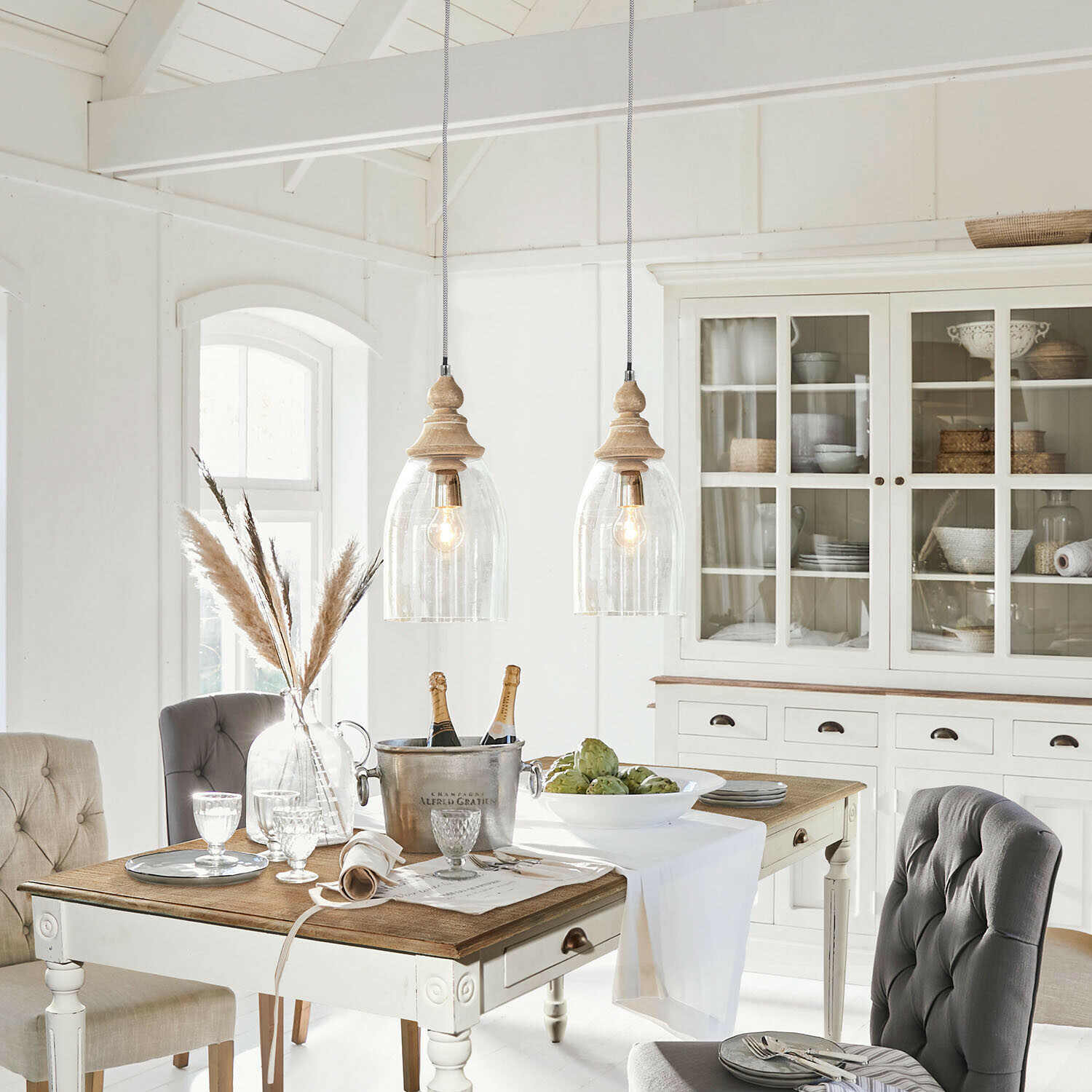 Suspension Raina Salle à manger rustique avec table en bois blanc, chaises rembourrées grises et buffet vitré décoré d’objets en osier