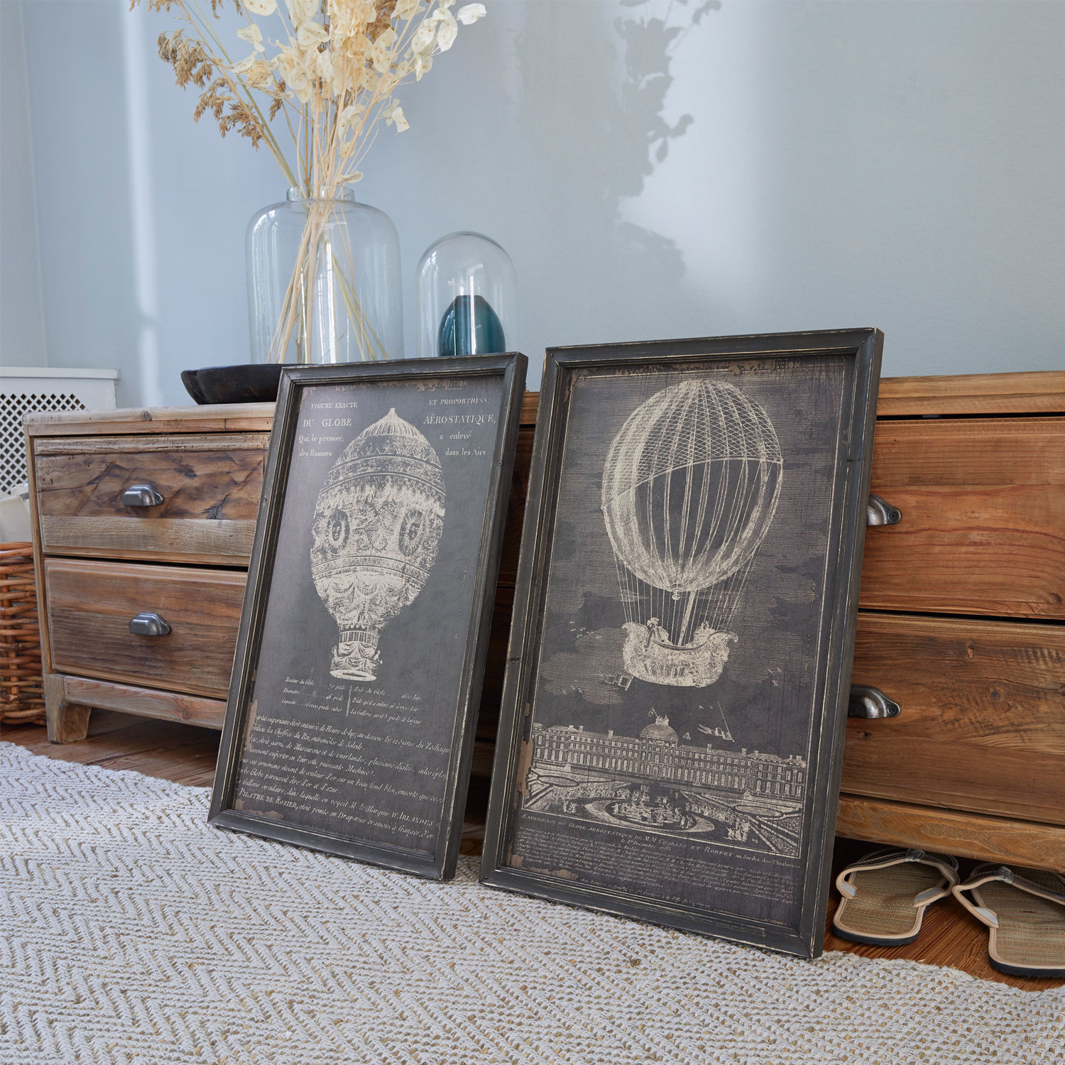 Lot de 2 tableaux Ortarsis Deux cadres noirs avec illustrations de montgolfières posés sur un sol en bois près d'une commode
