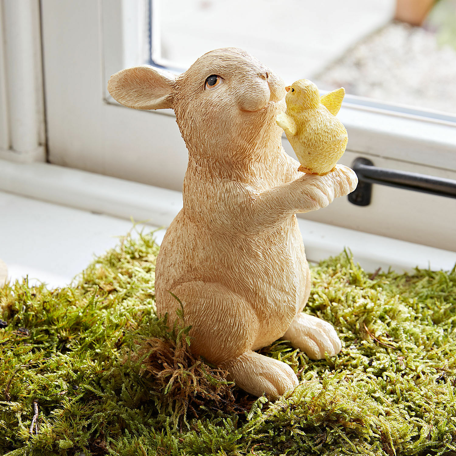 Lapin décoratif Raniah Figurine en résine d'un lapin beige tenant un poussin jaune sur de la mousse verte, décoration printanière