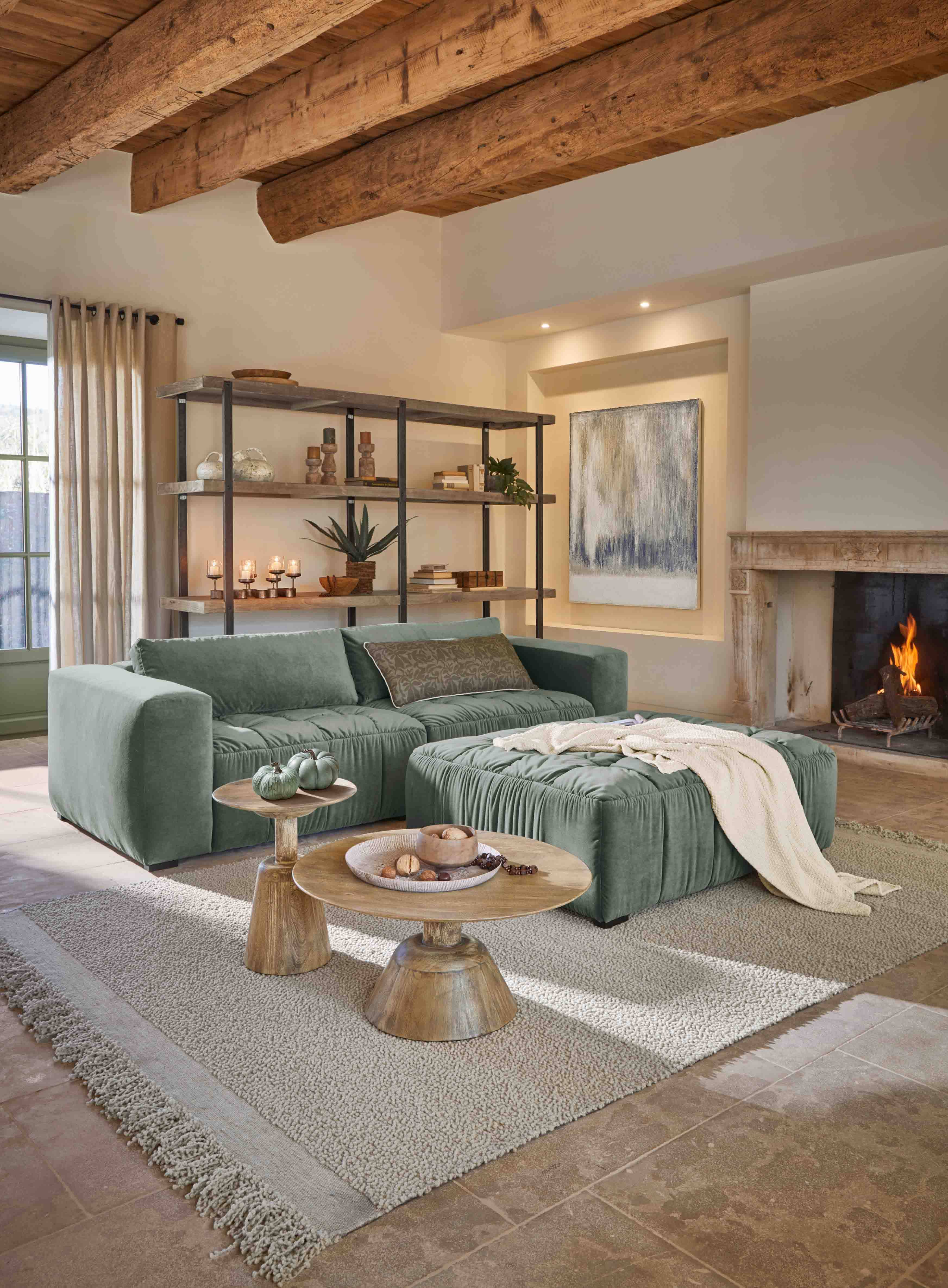 Salon chaleureux avec canapé vert en velours, pouf assorti, tables en bois et cheminée allumée