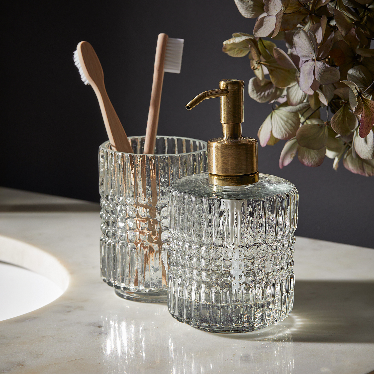 Distributeur de savon en verre avec pompe en laiton et gobelet en verre pour brosses à dents sur un comptoir en marbre