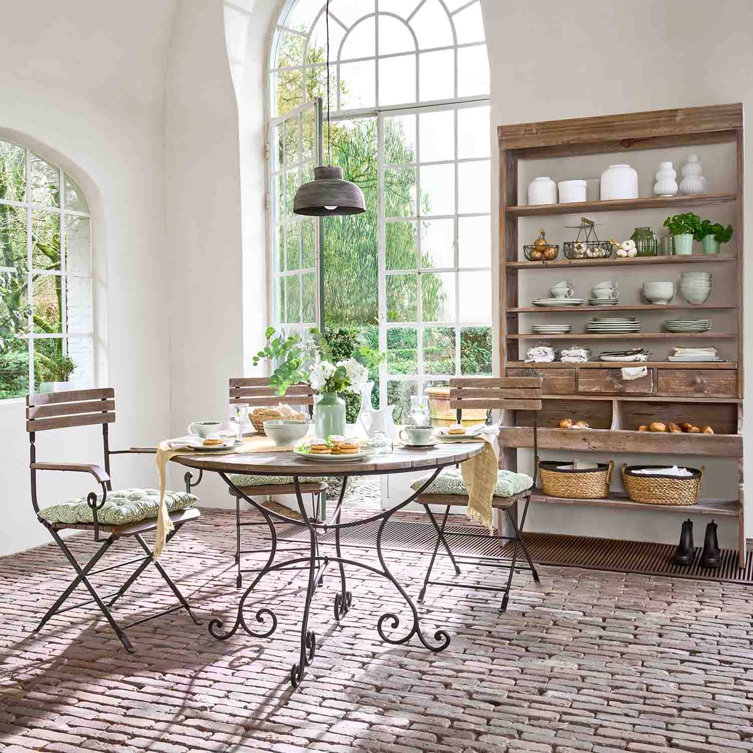 Salle à manger avec table ronde en métal et bois, chaises en bois, sol en briques et grande fenêtre