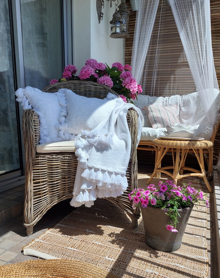 Balcon cosy avec fauteuil en osier, plaid blanc et fleurs roses en pot, ambiance décorative extérieure