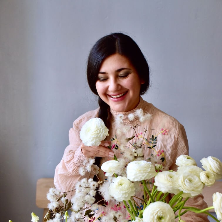 Femme souriante en blouse rose brodée tenant des fleurs blanches ranunculus dans une pièce lumineuse