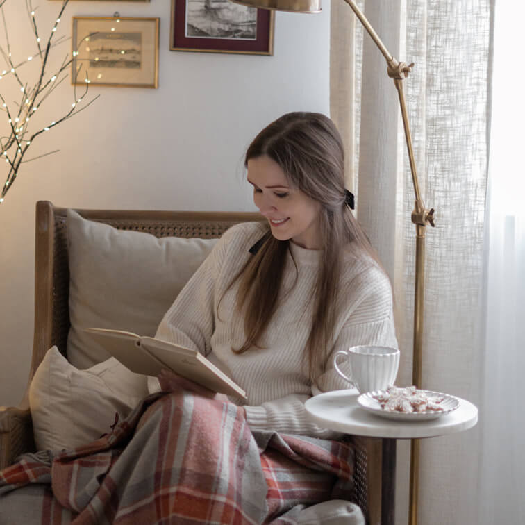 Femme lisant un livre sur un fauteuil en osier avec pull crème et couverture à carreaux chaude