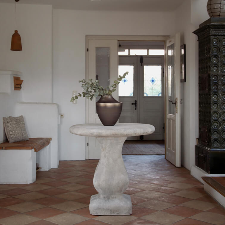 Table ronde en béton gris clair avec un vase en céramique foncée et des feuilles vertes dans un intérieur lumineux