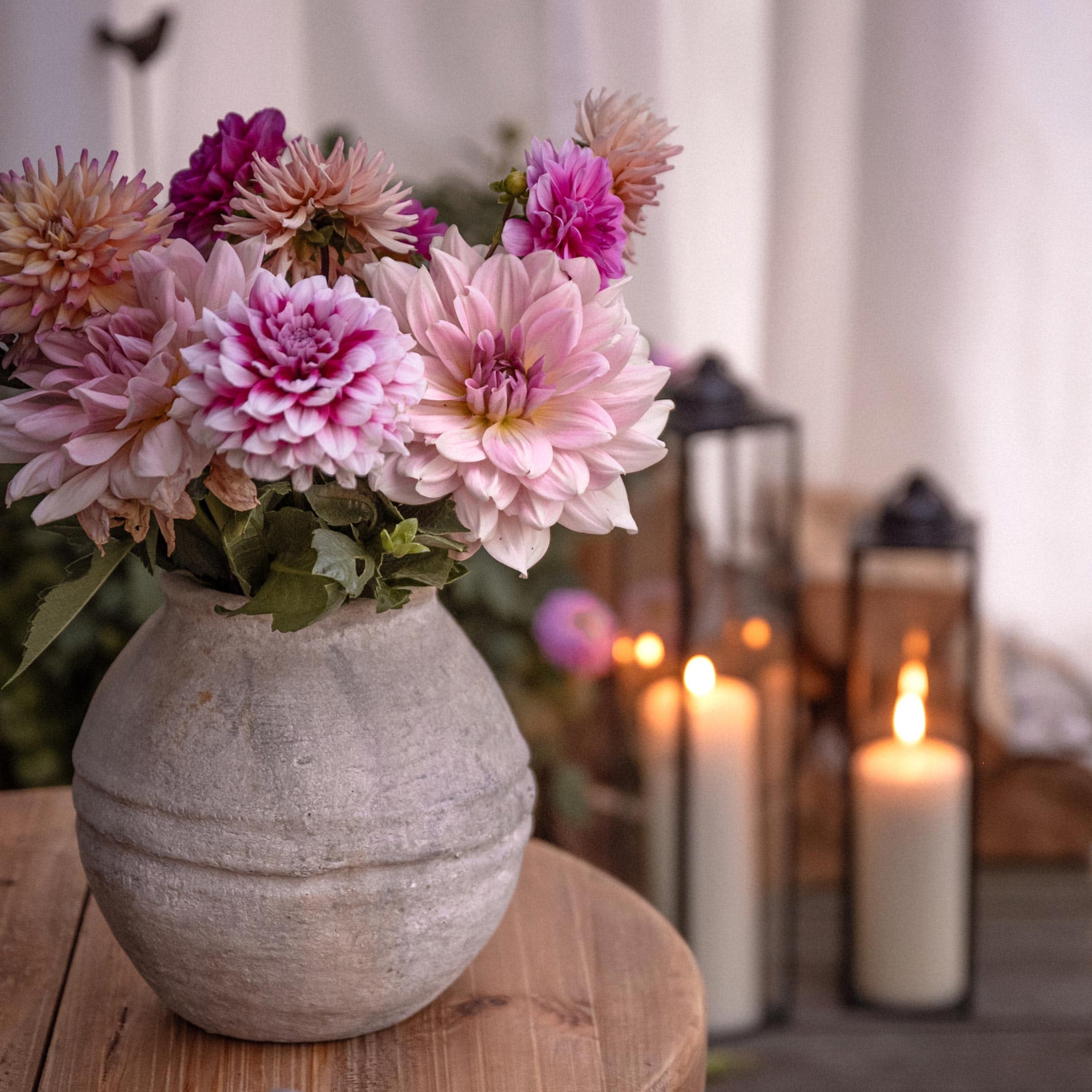 Vase en céramique grise avec fleurs roses et violettes sur table en bois, lanternes allumées en arrière-plan