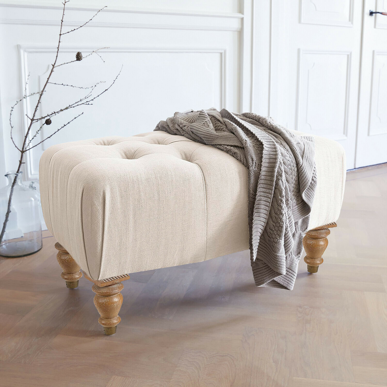Tabouret Durance Pouf beige rembourré avec pieds en bois et couverture tricotée grise dans un intérieur lumineux