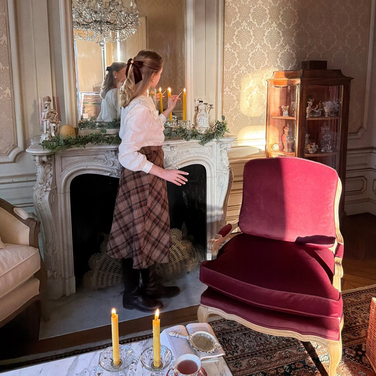 Femme allumant des bougies près d'une cheminée en marbre dans un salon élégant avec fauteuil bordeaux