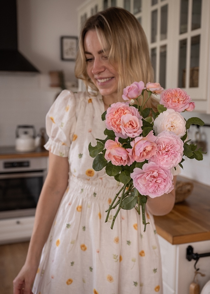 Femme en robe blanche à motifs floraux tenant un bouquet de roses roses dans une cuisine