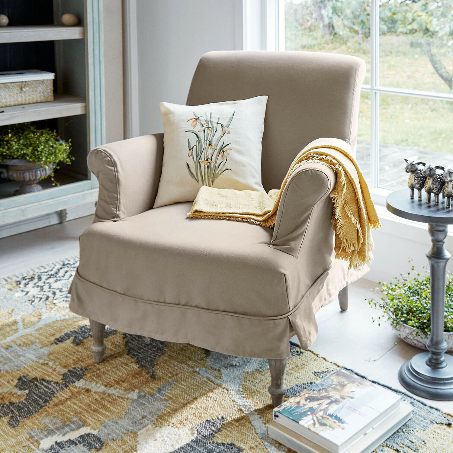 Fauteuil Everleighs Fauteuil beige en tissu avec coussin floral et plaid jaune dans un salon lumineux avec tapis coloré
