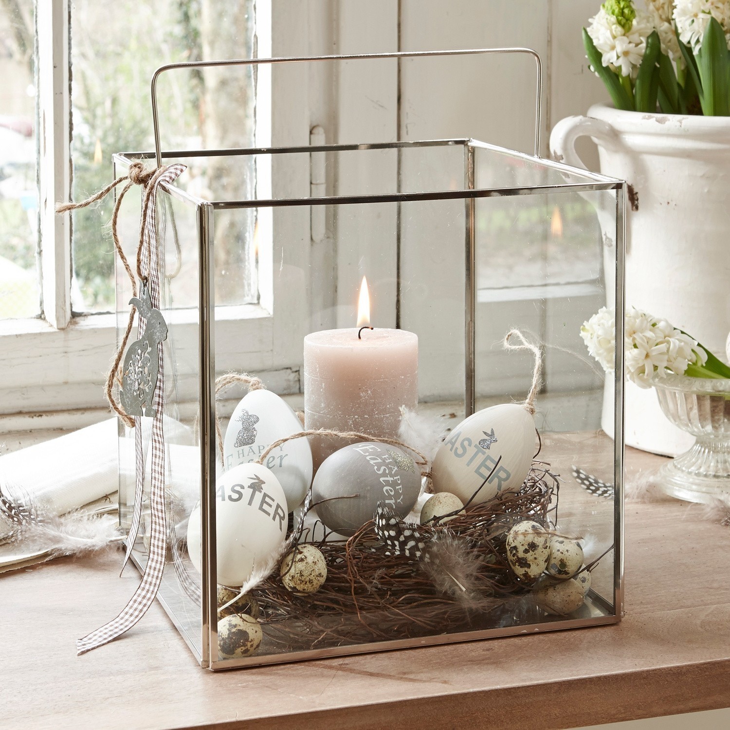 Photophore Otero Lanterne en verre avec cadre métallique, décorée d'œufs de Pâques et d'une bougie sur une table en bois