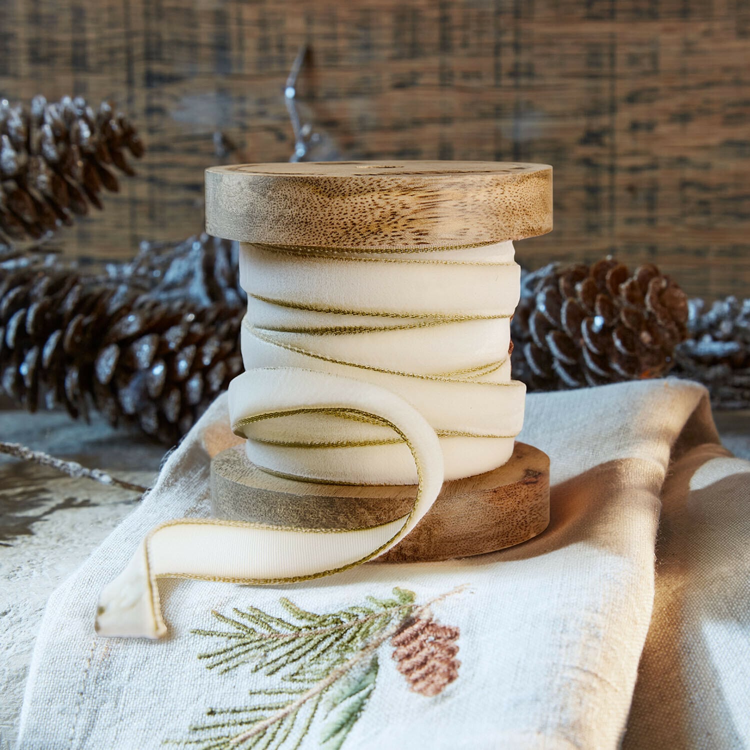 Ruban de velours Sorima Bobine en bois naturel avec ruban crème bordé de vert sur tissu en lin brodé d’un motif de pomme de pin