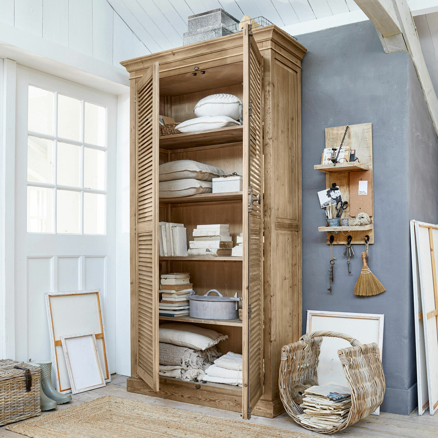 Armoire Farnham Armoire en bois naturel avec portes à persiennes, décorée de coussins et livres dans un intérieur lumineux