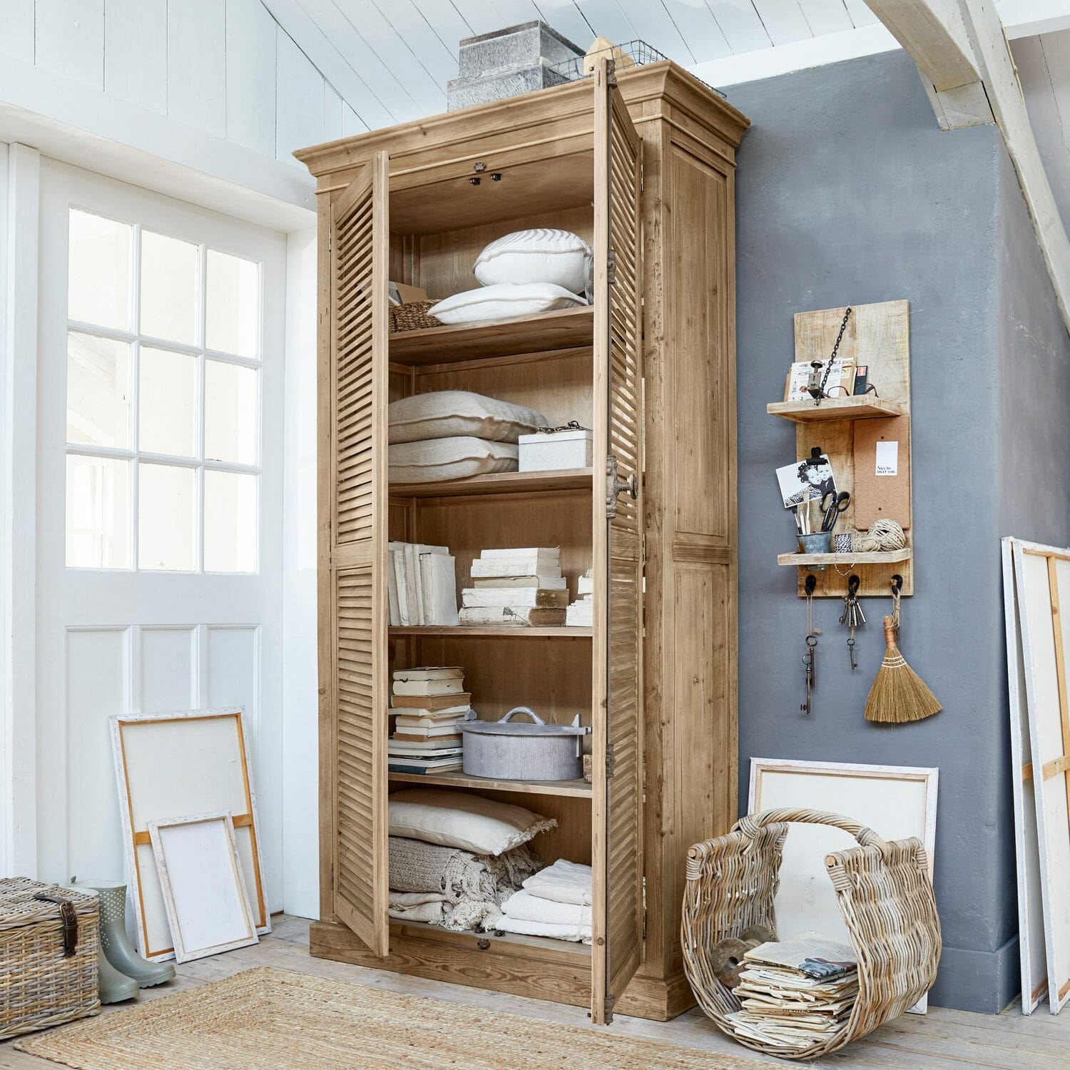 Armoire Farnham Armoire en bois naturel avec portes à persiennes, décorée de coussins et livres dans un intérieur lumineux