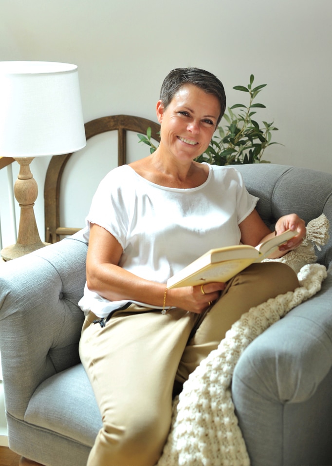 Femme souriante lisant un livre sur un canapé gris avec lampe et plante en arrière-plan