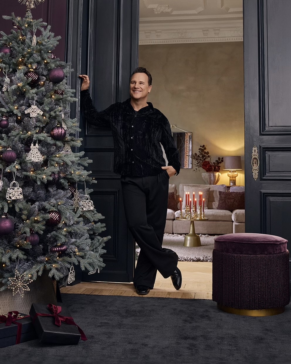 Homme en tenue noire près d'un sapin décoré de boules violettes dans un salon élégant avec bougies rouges allumées