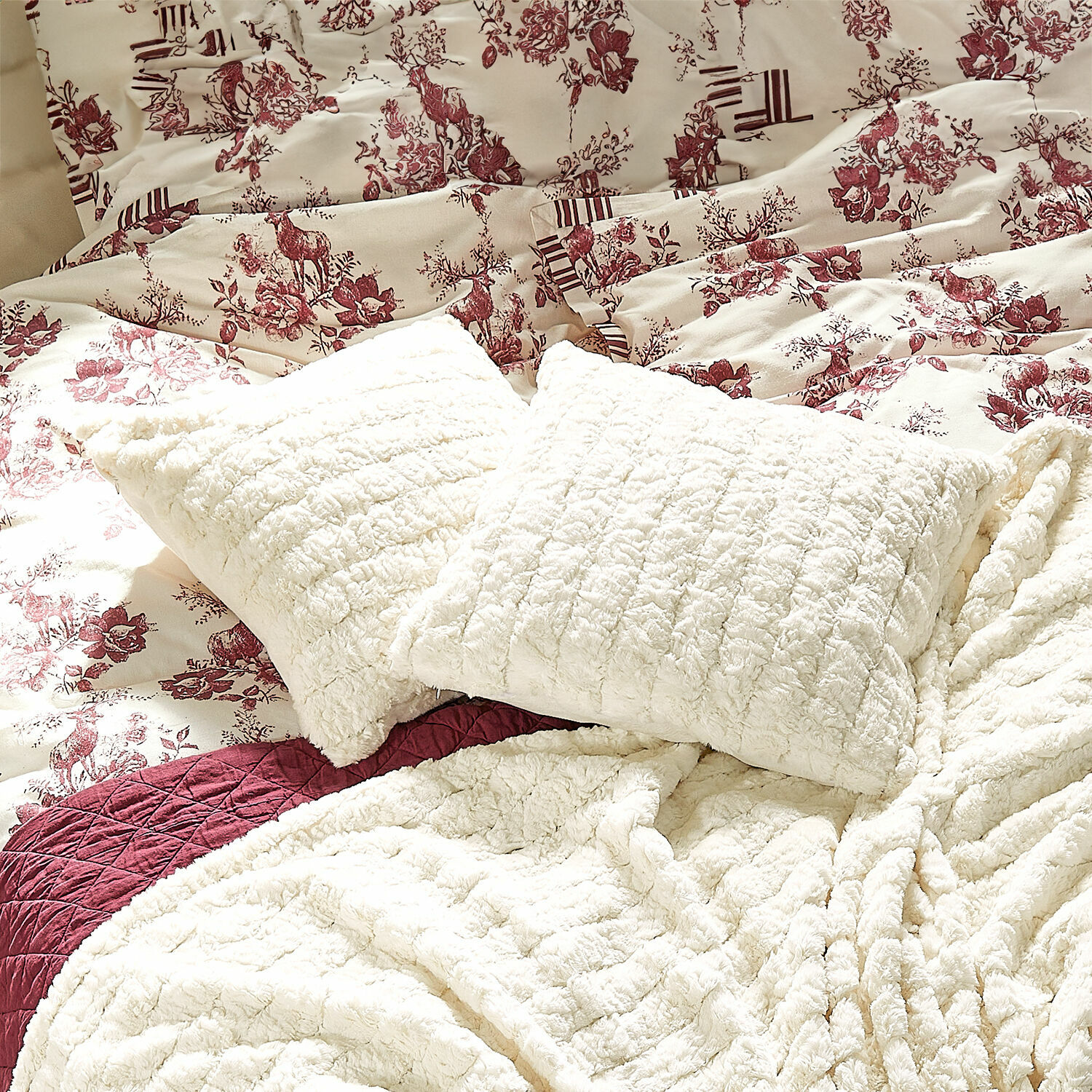 Housse de coussin Dorelice Coussins et couvertures en peluche crème sur un lit avec une literie à motifs floraux et cerfs bordeaux