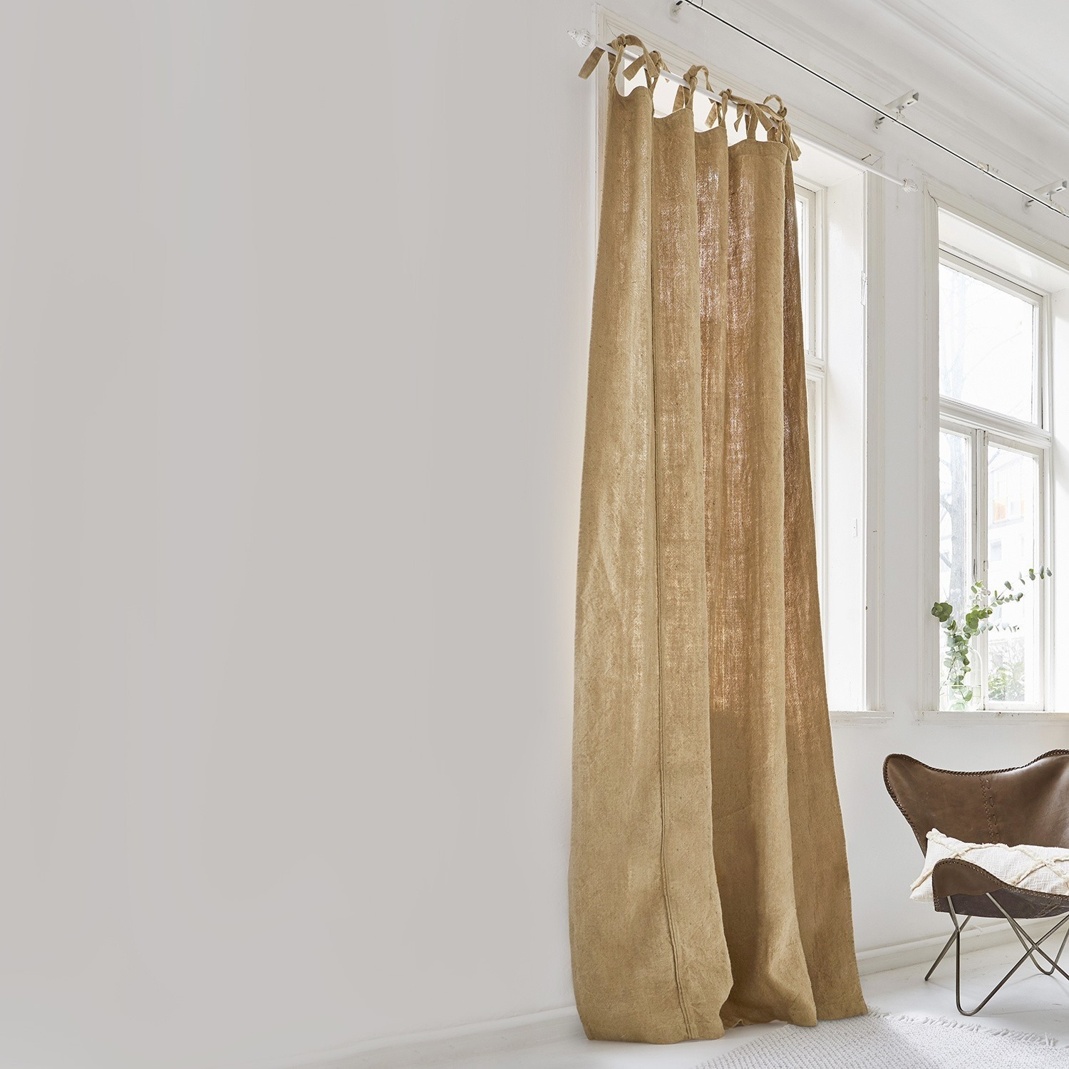 Rideau Uma Rideau beige en lin naturel dans une pièce lumineuse avec une grande fenêtre et un fauteuil marron