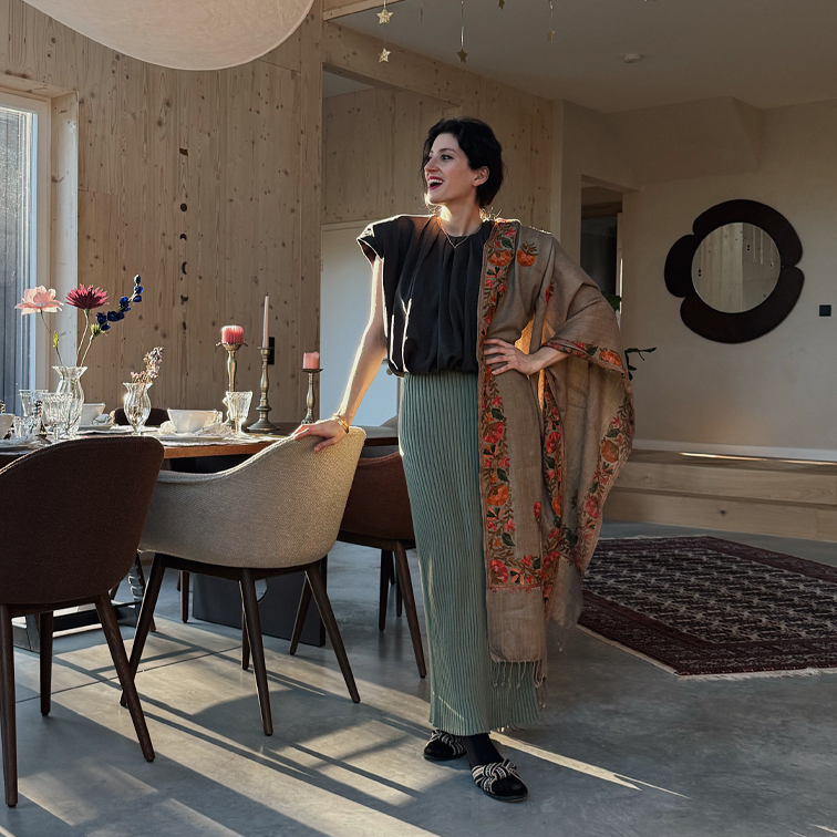Femme en jupe plissée verte et haut noir avec châle beige à motif floral dans un intérieur moderne