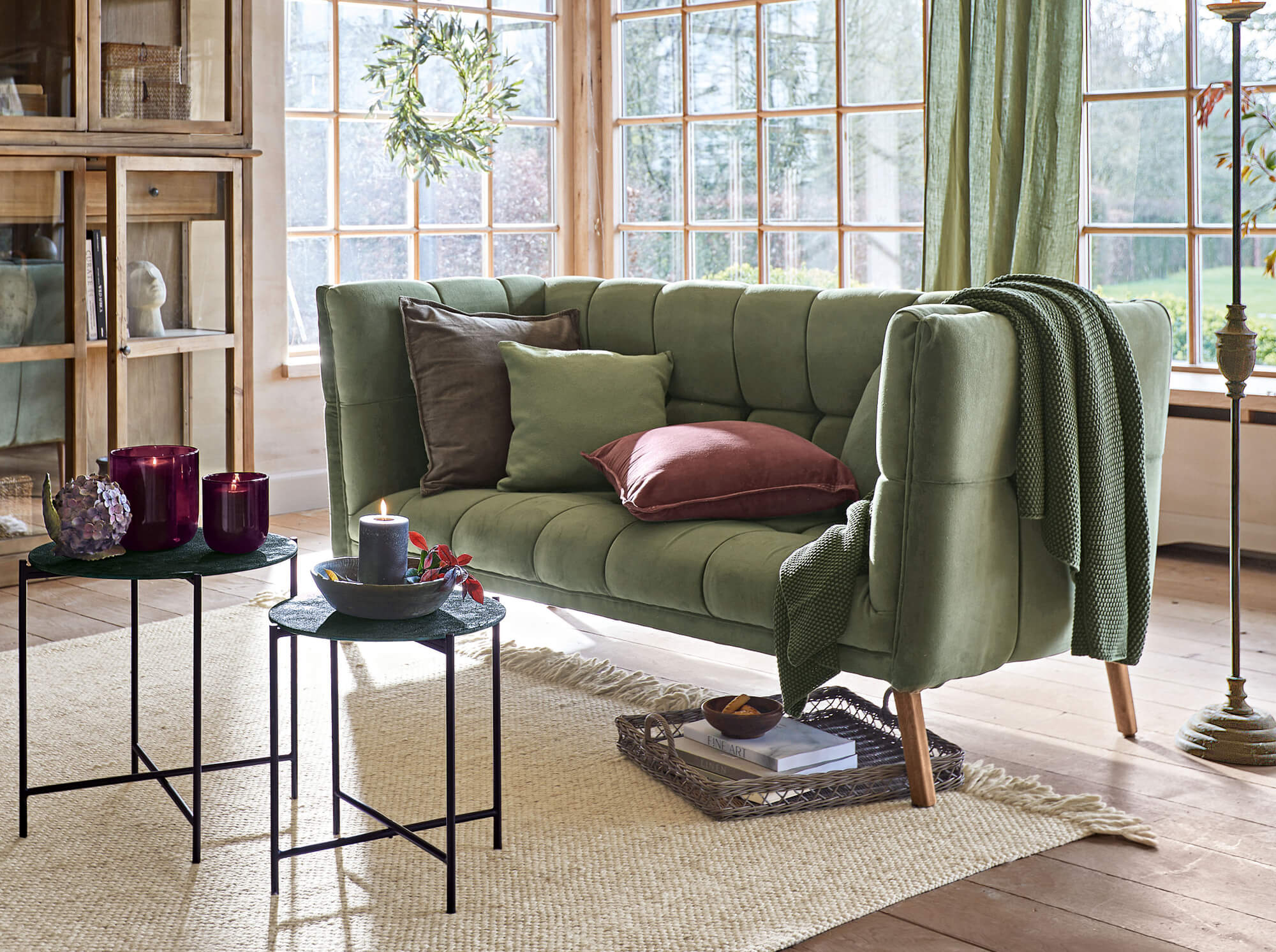 Canapé vintage vert en velours avec coussins et plaid dans un salon lumineux aux grandes fenêtres