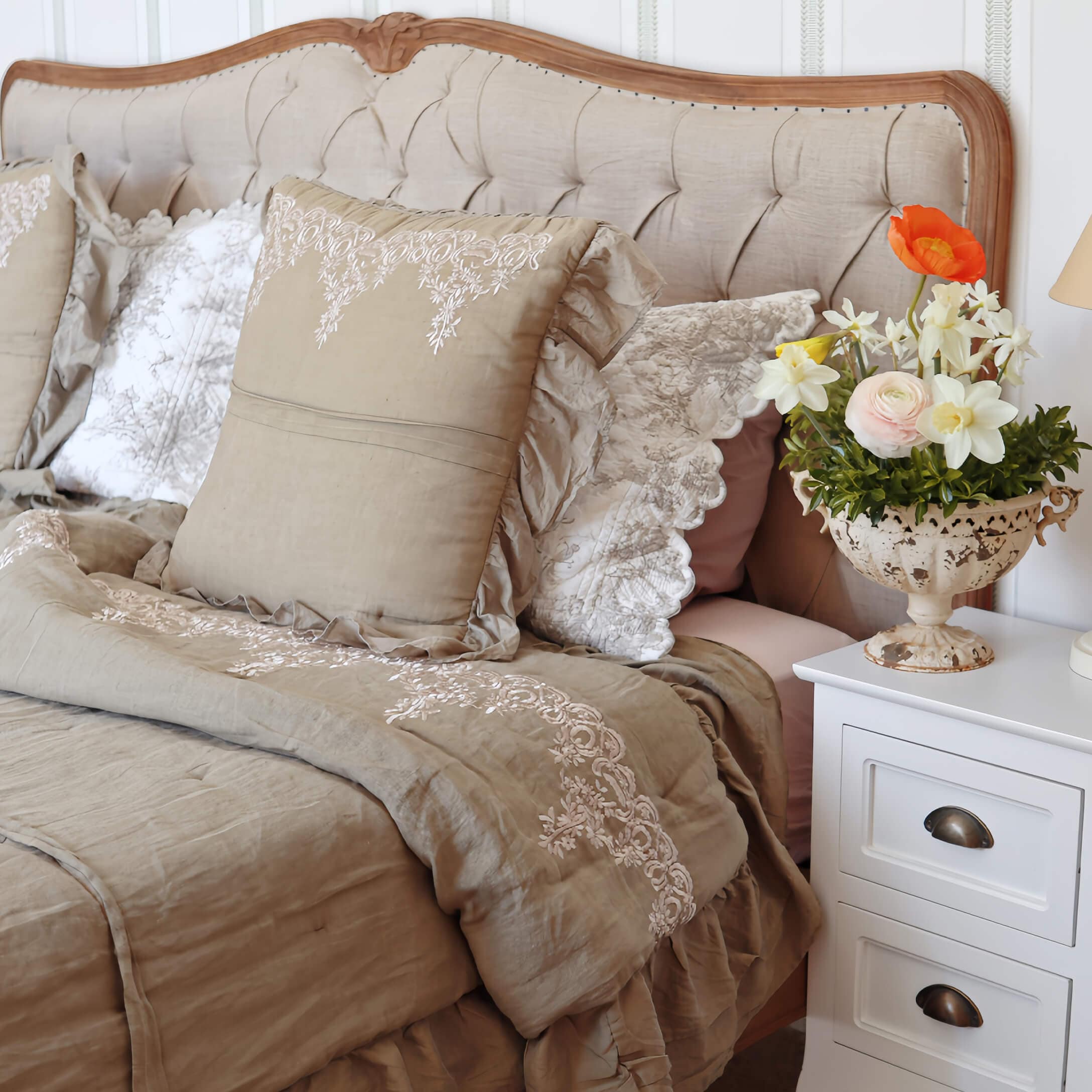 Linge de lit beige avec coussins brodés et couverture sur un lit à tête de lit capitonnée