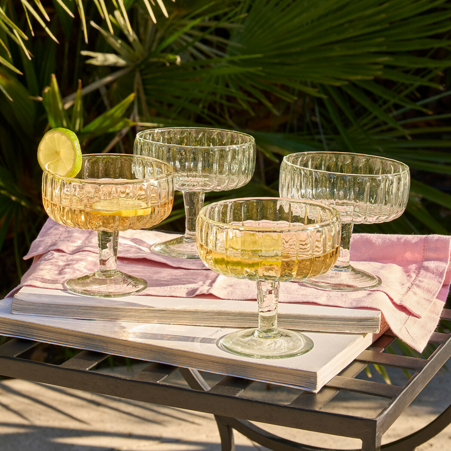 Ensemble de quatre verres à cocktail en verre texturé transparent sur une table en métal avec livre et serviette rose