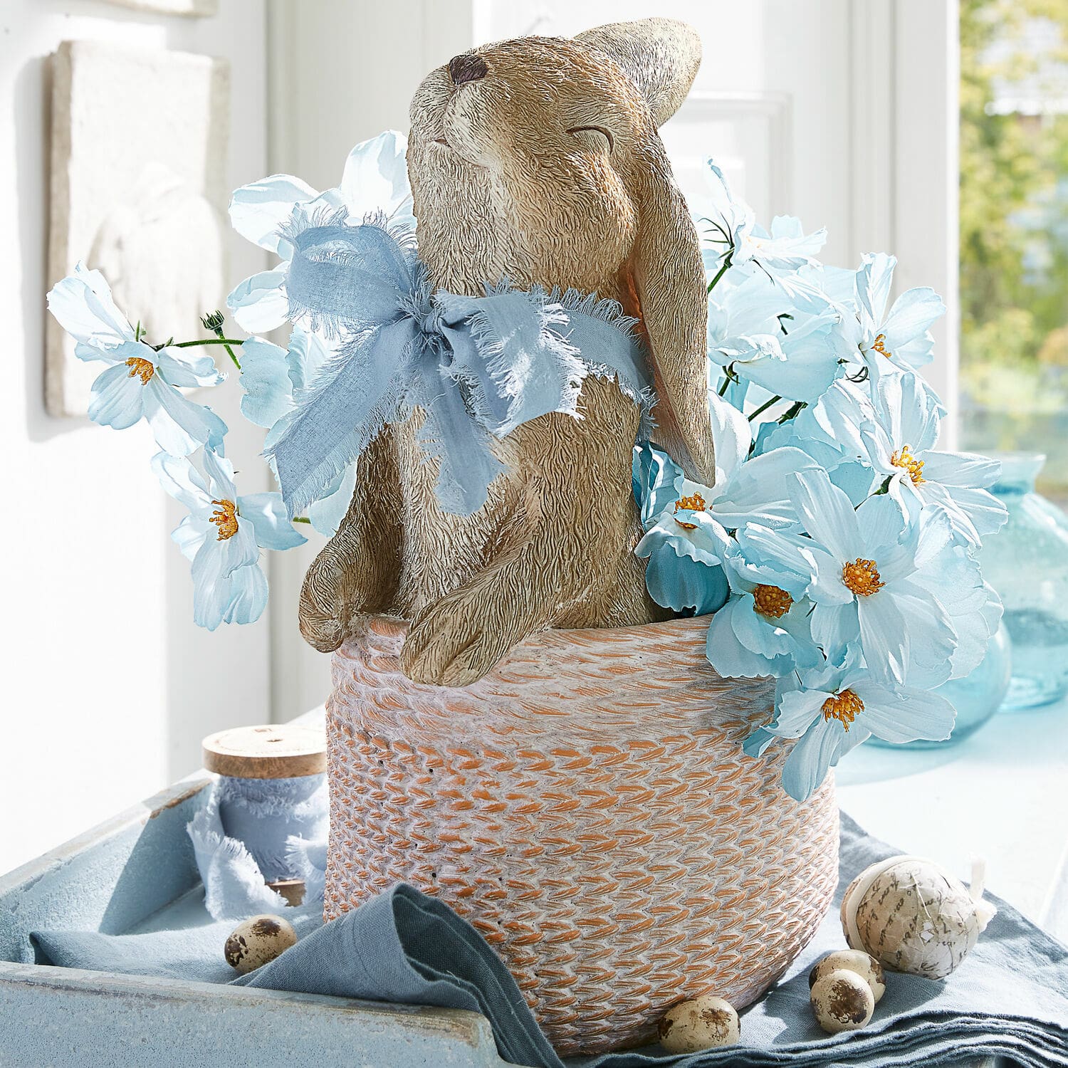 Cache-pot Merisette Lapin décoratif en matériau dans un panier avec fleurs bleues et ruban pour décoration printanière