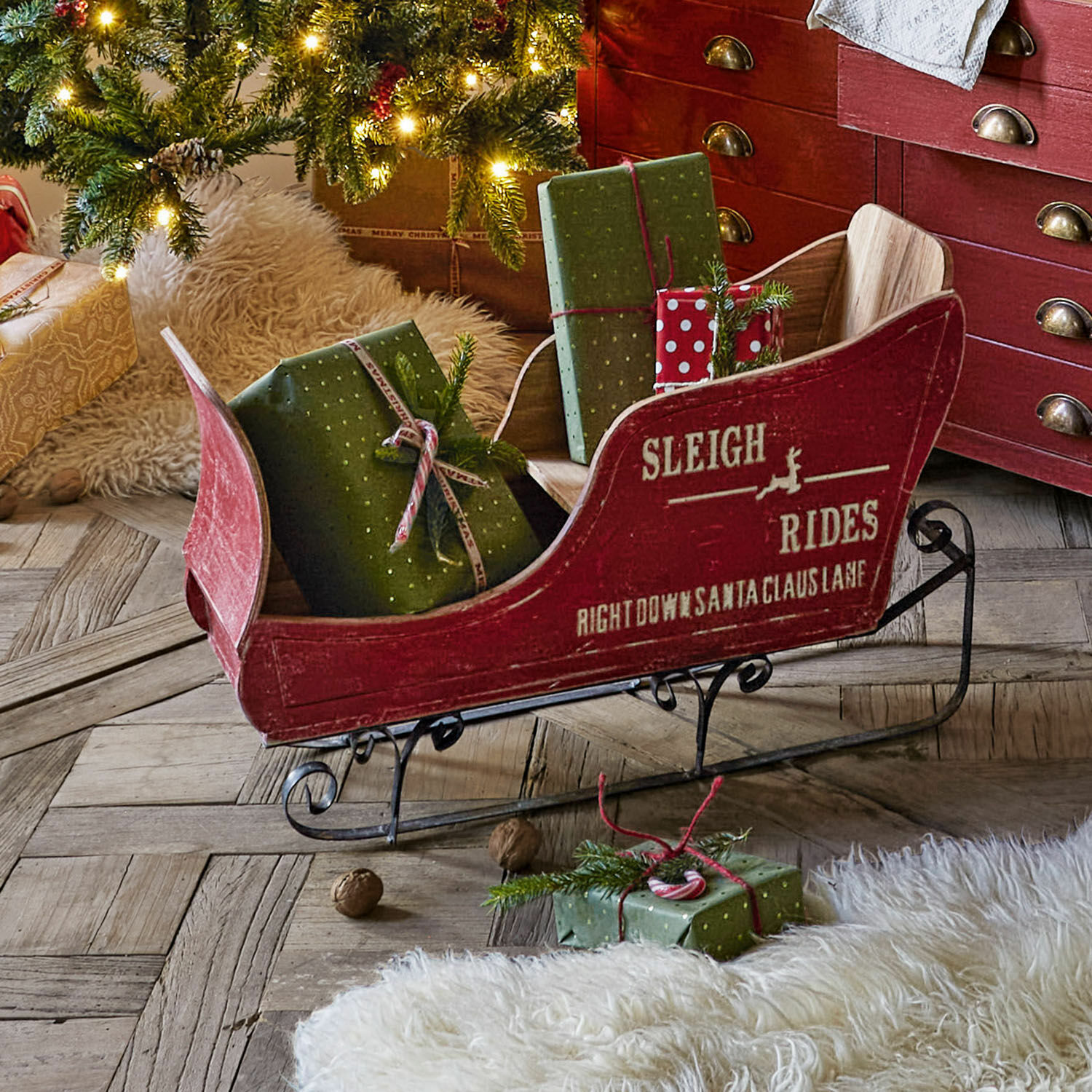 Luge décorative Maélys Luge en bois rouge avec patins en métal et cadeaux verts sous un sapin décoré sur un sol en parquet