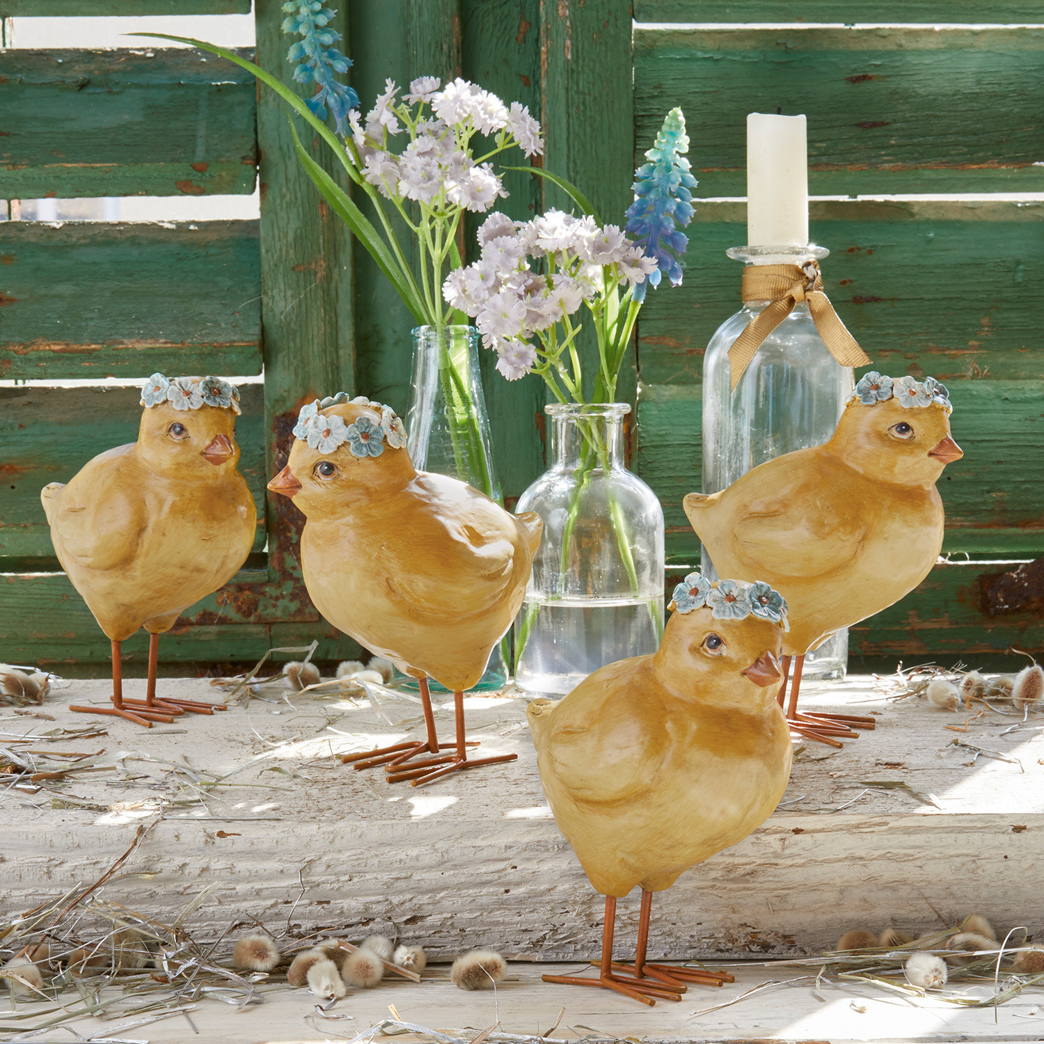 Lot de 4 poussins Dorette Quatre poussins décoratifs jaunes en matériau avec couronnes de fleurs bleues sur fond de bois vert vieilli