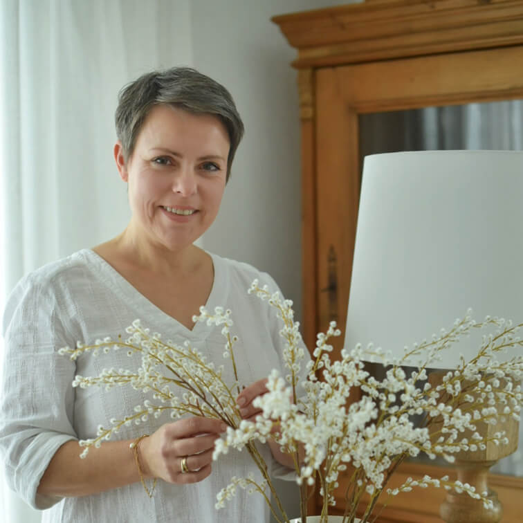 Femme en chemise blanche tenant des branches décoratives à fleurs blanches dans un intérieur lumineux