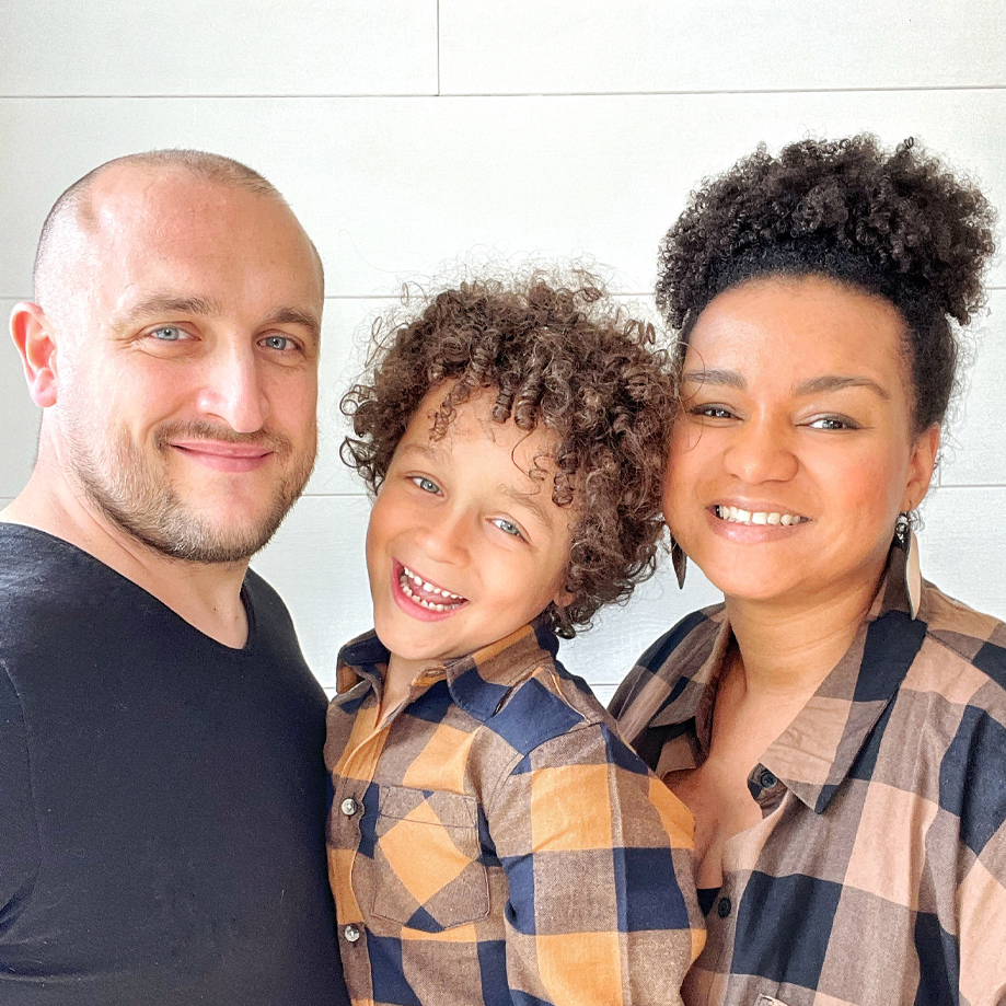 Famille souriante devant un mur clair, enfant et adultes en vêtements décontractés.