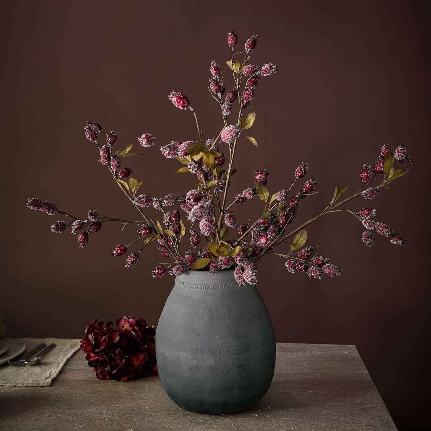 Vase en céramique grise avec branches décoratives aux fruits rouges et feuilles vertes sur table en bois
