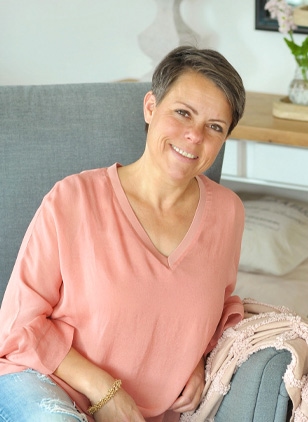 Femme souriante en haut rose assise sur un canapé gris dans un intérieur lumineux