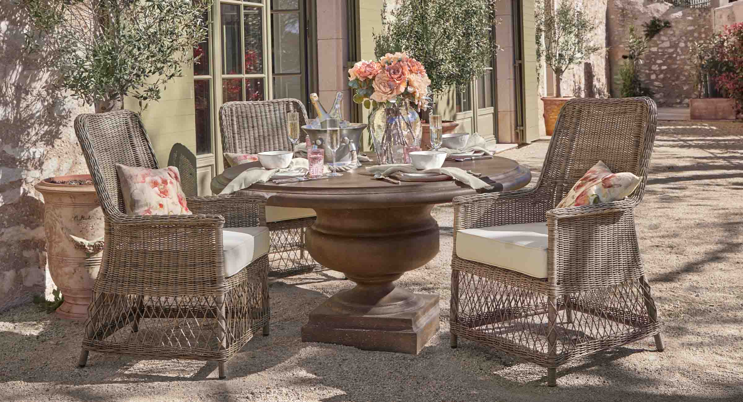 Table ronde en bois brun avec quatre chaises en rotin et coussins clairs décorée de fleurs en extérieur